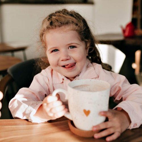 Indoor close-up photo og little charming girl with blond curly hair has fun and drinking cacao with happy smile in cafe and holding a big cup